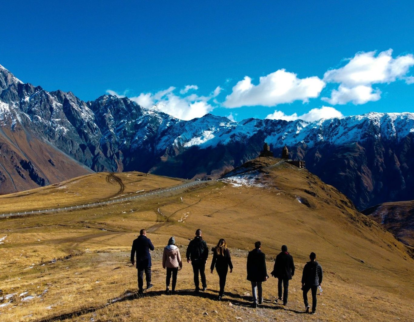 Groupe de six personnes marchant sur un sentier de montagne avec des sommets enneigés en arrière-plan.