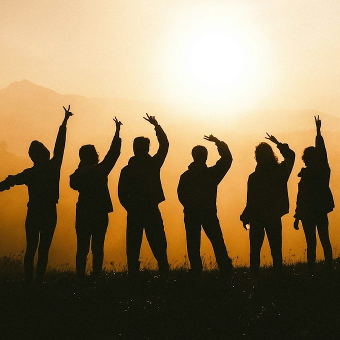 Silhouette de six personnes levant les bras au coucher de soleil en montagne.